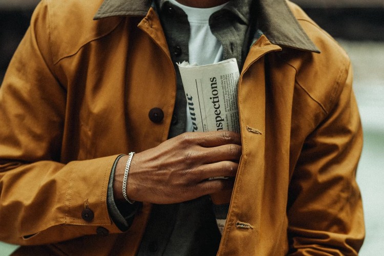 A person in a mustard yellow jacket with a dark collar holds a folded newspaper in their left hand.