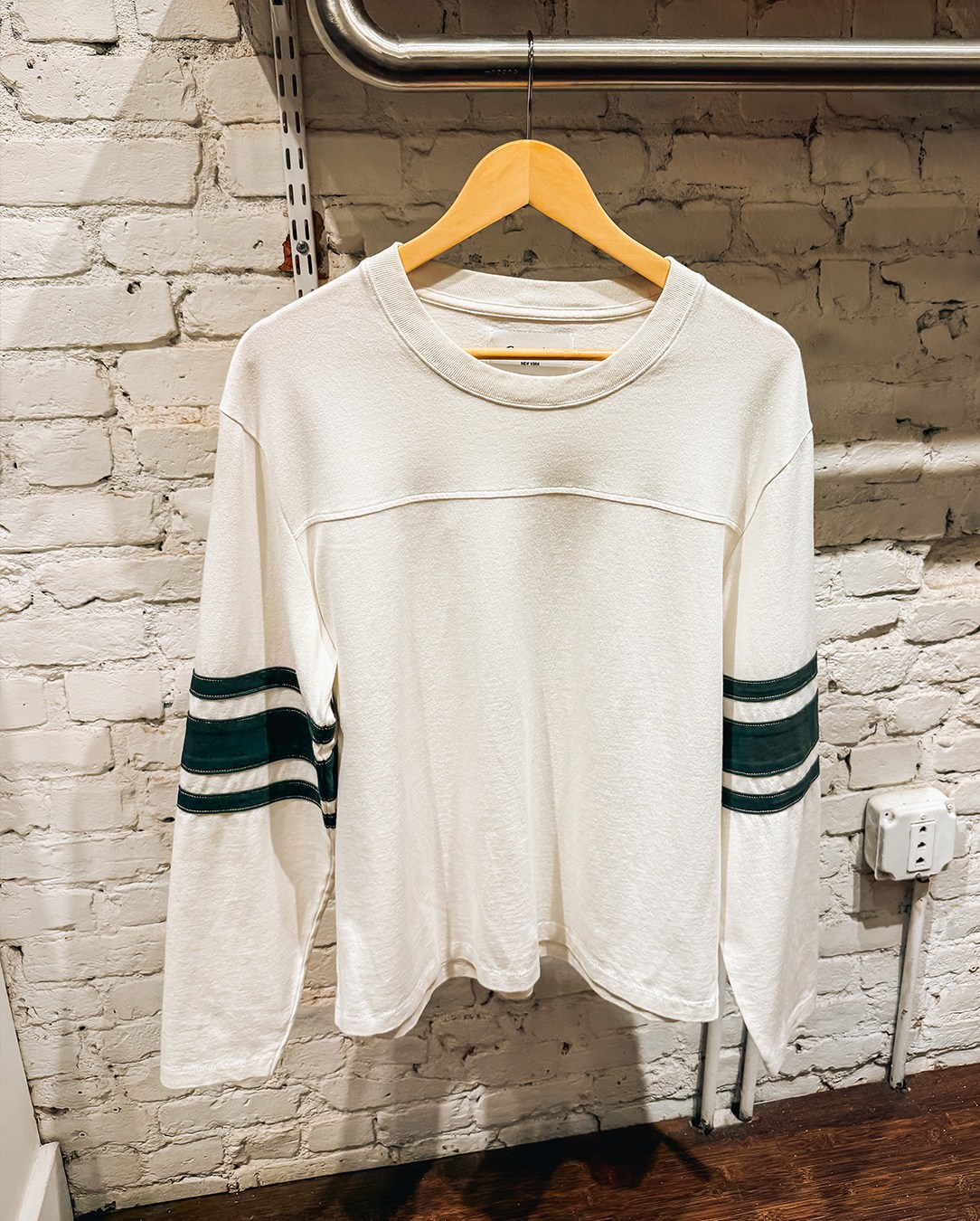 White long-sleeved shirt with green stripes on the sleeves, hanging on a wooden hanger against a brick wall.