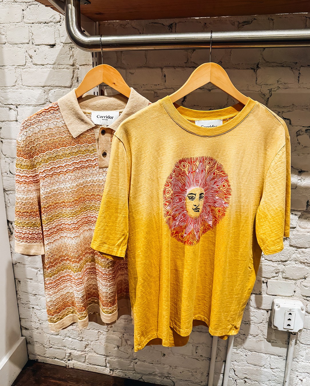 Two shirts on hangers against a white brick wall: a beige polo with orange and green waves, and a yellow tee with a red sun face.