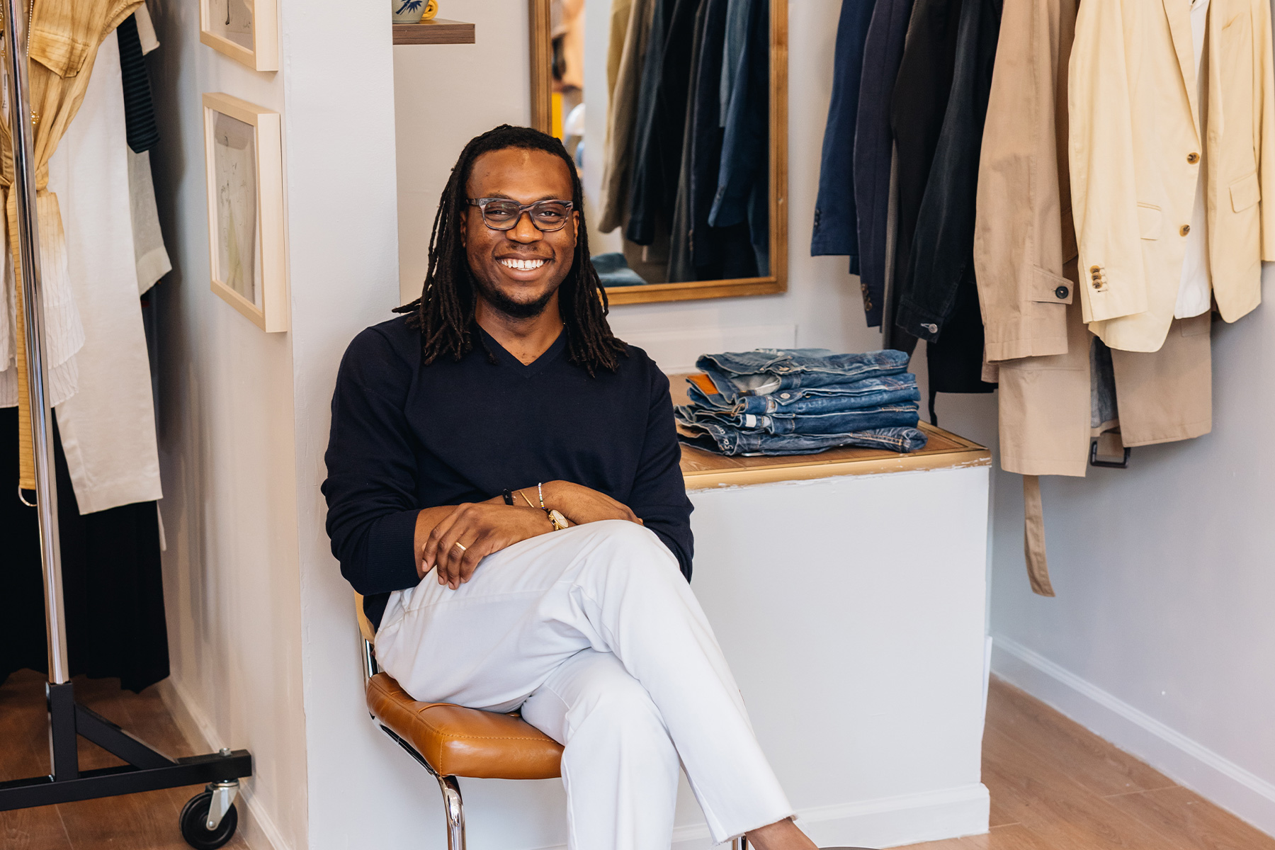 Daniel Dele-Tunji, wearing a navy sweater and white pants, sitting in a clothing store.