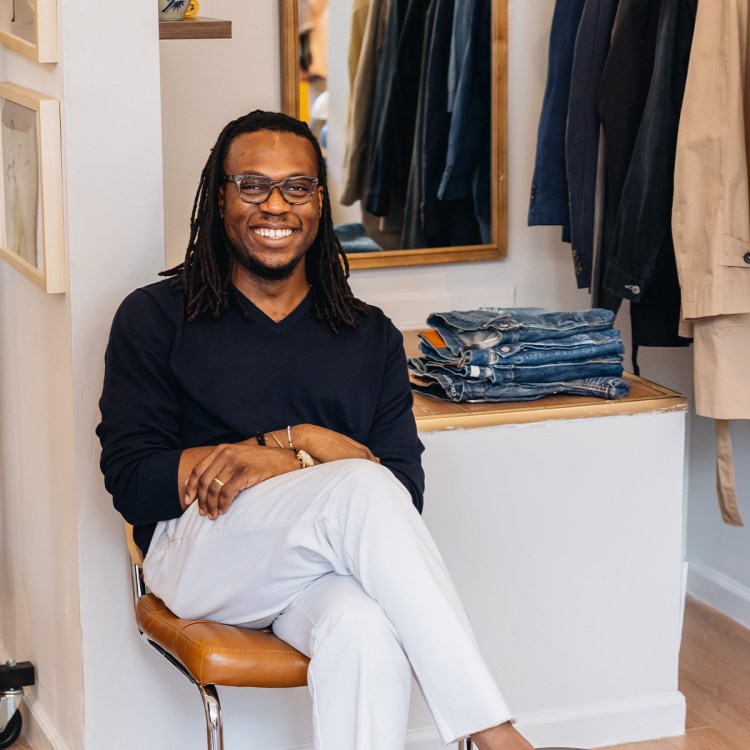 Daniel Dele-Tunji, wearing a navy sweater and white pants, sitting in a clothing store.