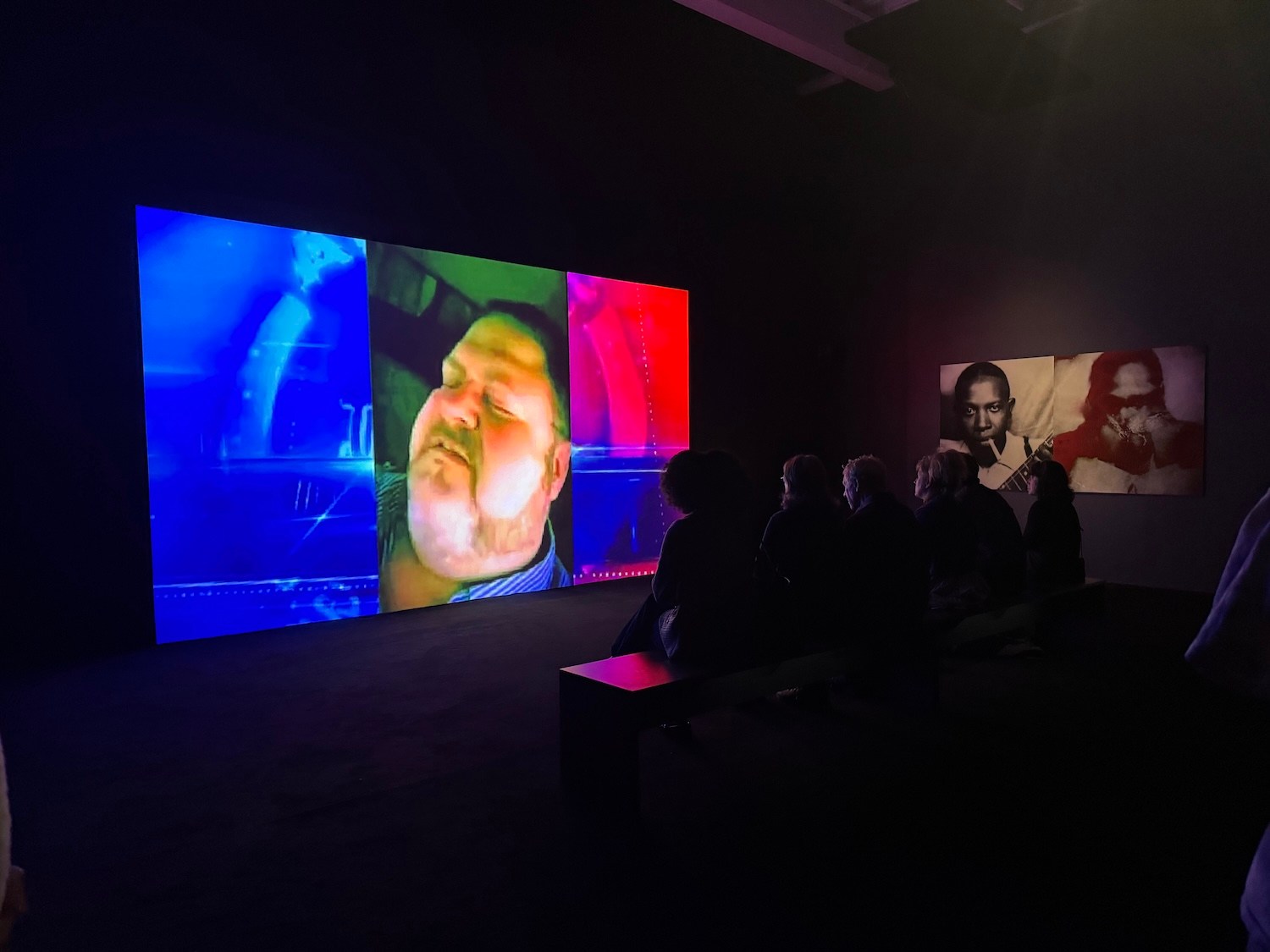 People watch a large video screen showing a man's face, with other art on the wall in a dimly lit room.