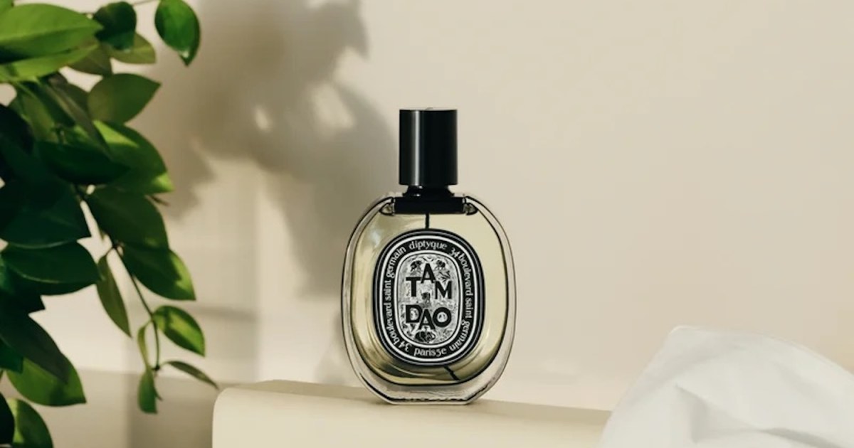 Diptyque Tam Dao perfume bottle with a black cap and label, light liquid, next to green leaves and a white cloth.