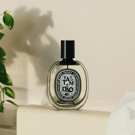 Diptyque Tam Dao perfume bottle with a black cap and label, light liquid, next to green leaves and a white cloth.