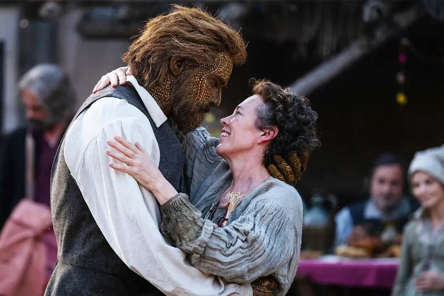 A woman with curly hair smiles up at a man with a hairy, woven face, embracing him.