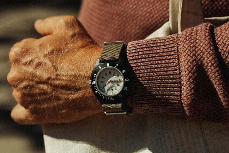 A person's arm with a black watch, white face, and olive green strap, wearing a rust-colored sweater.