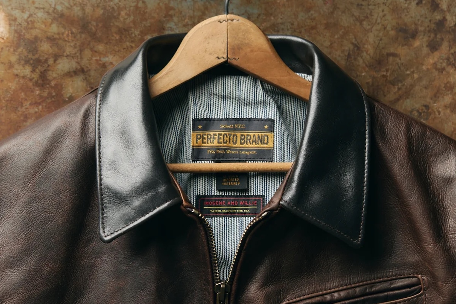 Man in a brown leather jacket, light blue denim shirt, and olive pants with a patterned belt.