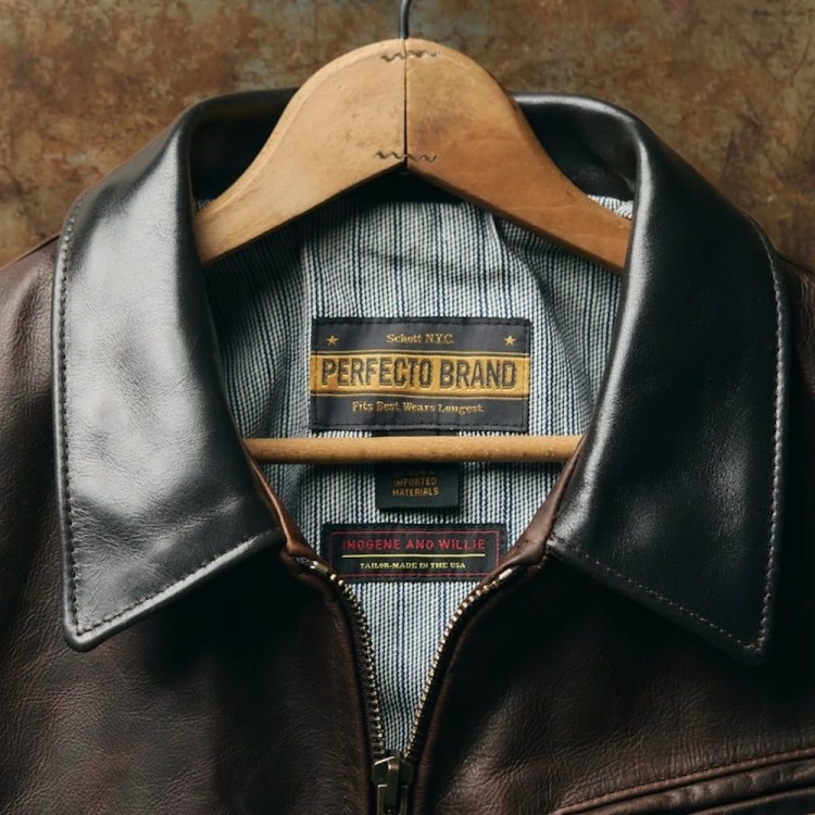 Man in a brown leather jacket, light blue denim shirt, and olive pants with a patterned belt.