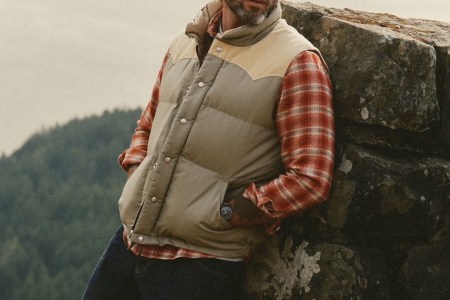 Man in a beige puffer vest with tan shoulders, red plaid shirt, and jeans, leaning against a rock wall.