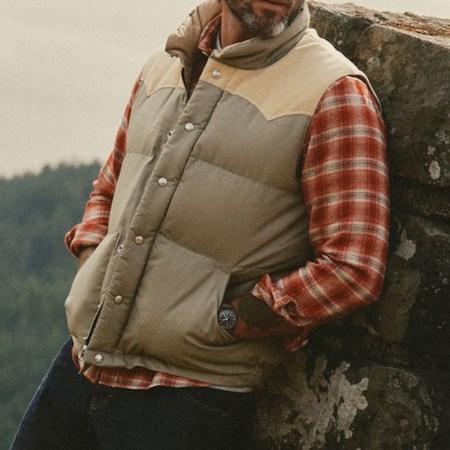 Man in a beige puffer vest with tan shoulders, red plaid shirt, and jeans, leaning against a rock wall.