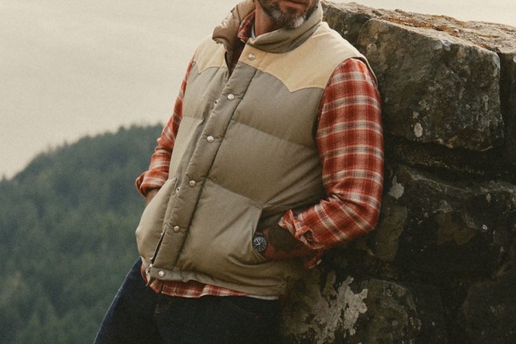 Man in a beige puffer vest with tan shoulders, red plaid shirt, and jeans, leaning against a rock wall.