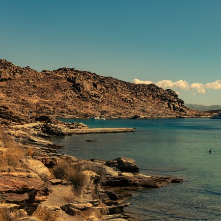 Rocky coastline with a dirt path, a small pier, boats in turquoise water, and people swimming.