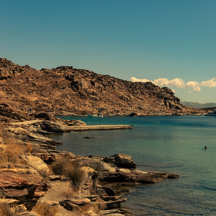 Rocky coastline with a dirt path, a small pier, boats in turquoise water, and people swimming.