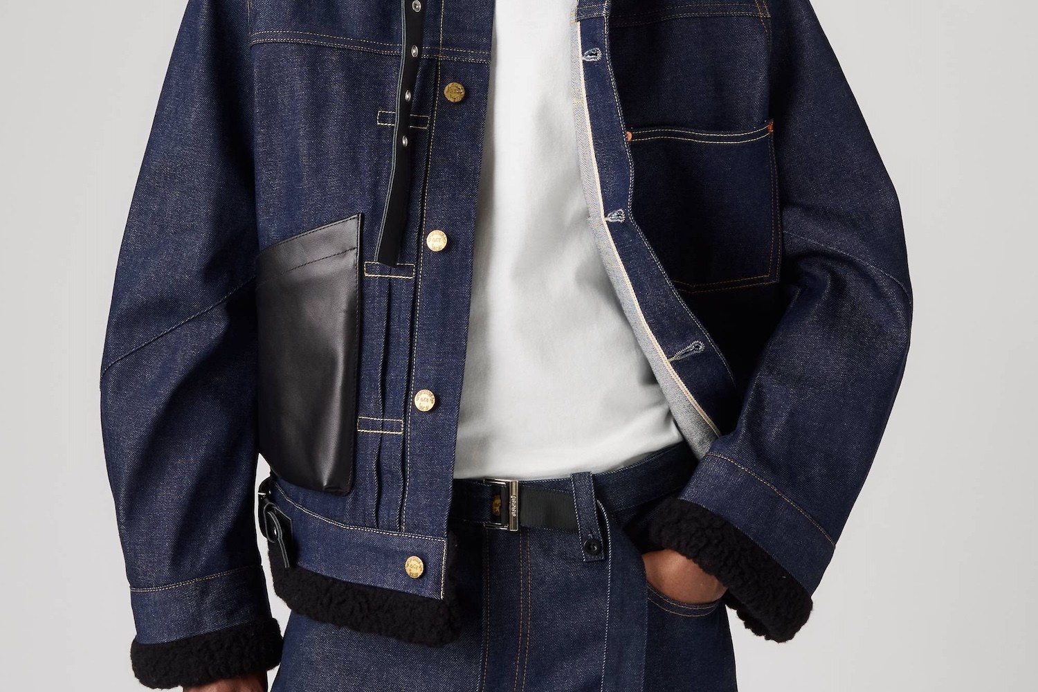 Person wearing a dark denim jacket with gold buttons, a black leather pocket, and black fleece trim, over a white t-shirt.