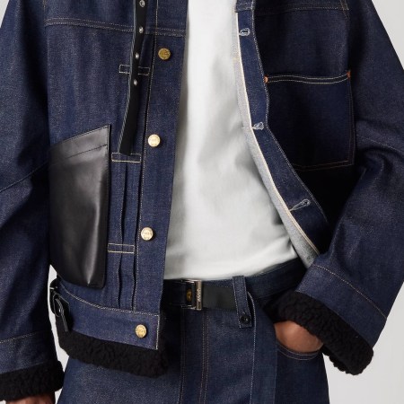Person wearing a dark denim jacket with gold buttons, a black leather pocket, and black fleece trim, over a white t-shirt.