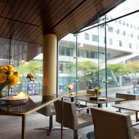 Modern restaurant interior with floor-to-ceiling windows overlooking a city street, tables set with yellow flowers.
