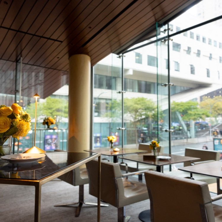 Modern restaurant interior with floor-to-ceiling windows overlooking a city street, tables set with yellow flowers.