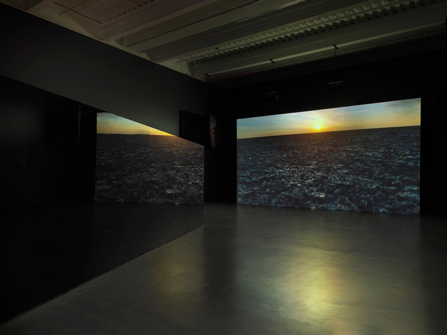 Two large screens in a dark room display a sunset over a choppy ocean, with reflections on the floor.