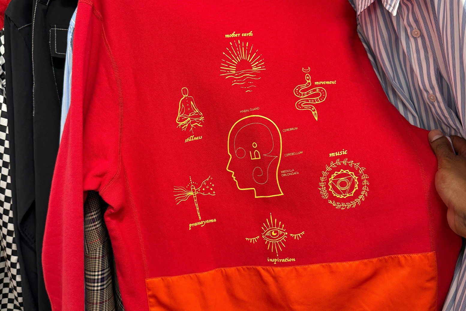 Red sweatshirt with orange bottom panel and gold line art of a head, snake, sun, and other symbols.