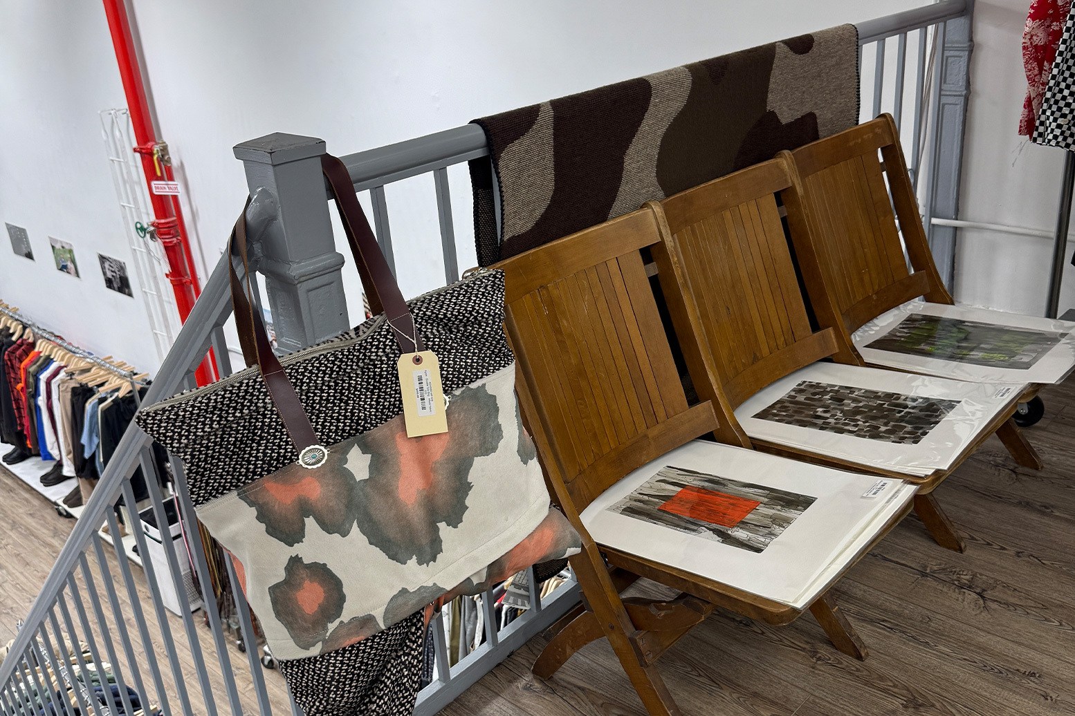 A tote bag with an abstract orange and black pattern hangs on a gray railing next to three wooden chairs with art prints.