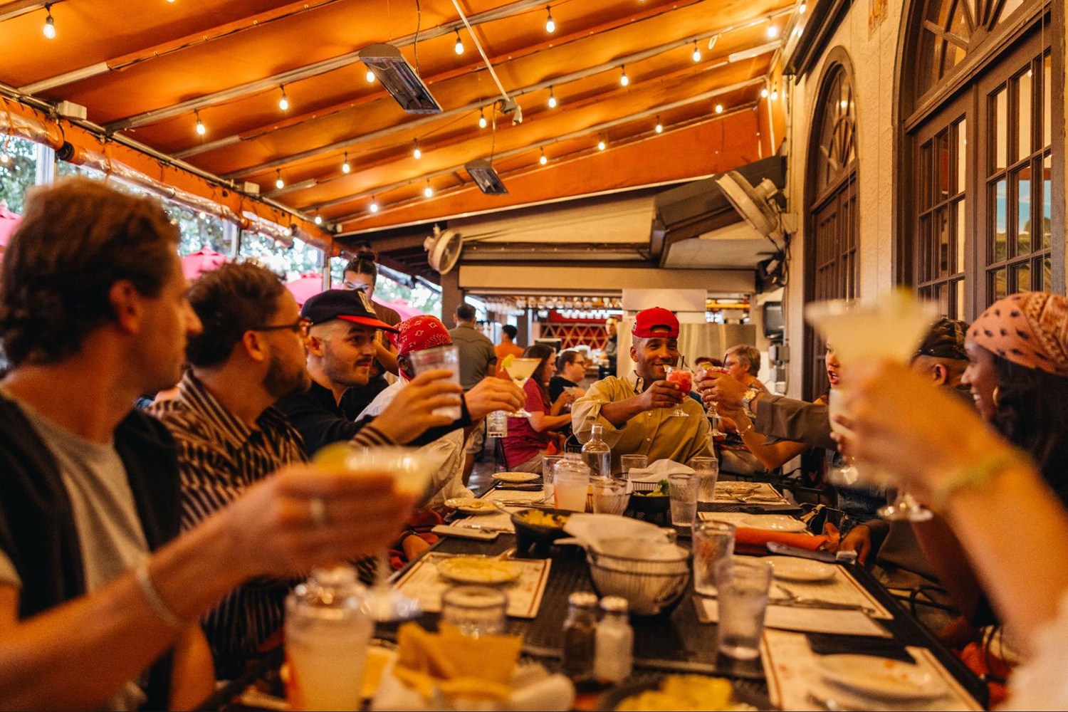 Friends at an outdoor restaurant raise their drinks in a toast under an orange awning with string lights.