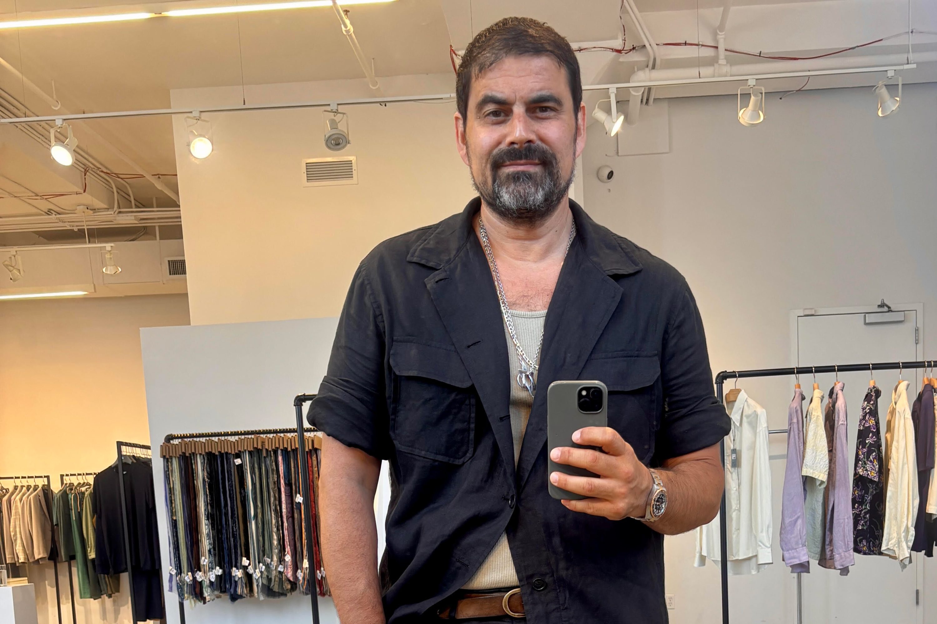 Man with dark hair and beard, wearing a black shirt and silver necklace, takes a mirror selfie in a clothing store.