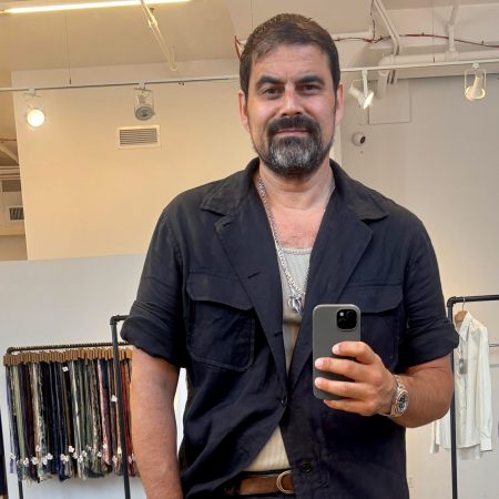 Man with dark hair and beard, wearing a black shirt and silver necklace, takes a mirror selfie in a clothing store.