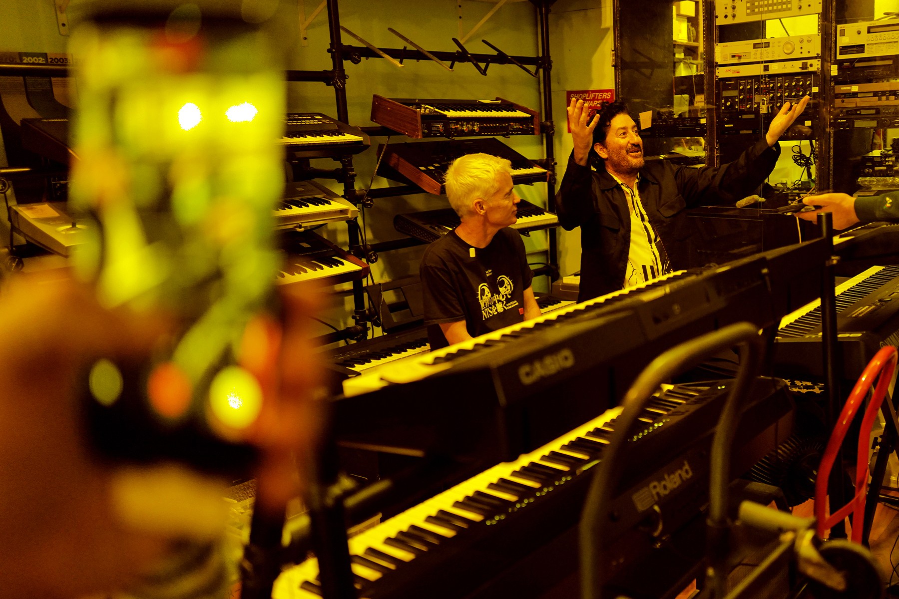 A person filming two men in a studio filled with keyboards and music equipment. One man gestures, the other watches.
