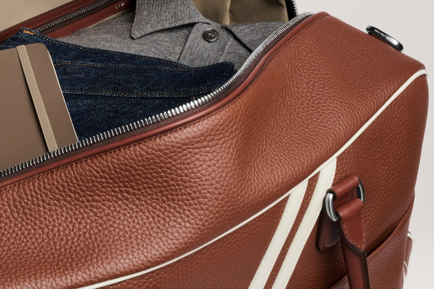 Open brown leather duffel bag with white stripes, revealing a notebook, jeans, and a gray polo shirt inside.
