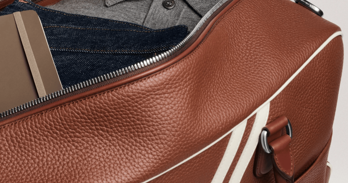 Open brown leather duffel bag with white stripes, revealing a notebook, jeans, and a gray polo shirt inside.