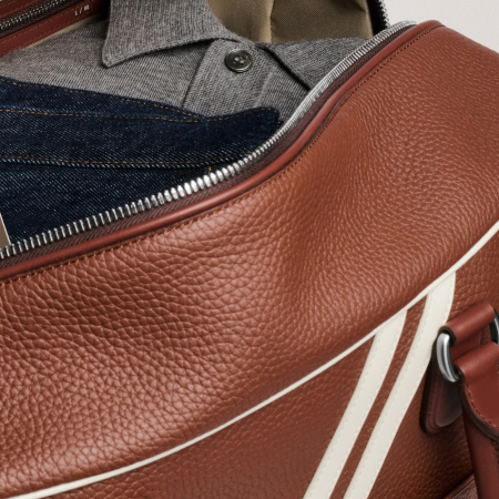 Open brown leather duffel bag with white stripes, revealing a notebook, jeans, and a gray polo shirt inside.