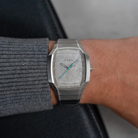 A person's arm wearing a silver Wren watch with a light gray, textured face and teal second hand.