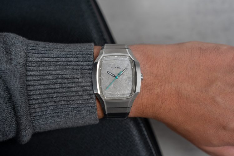 A person's arm wearing a silver Wren watch with a light gray, textured face and teal second hand.