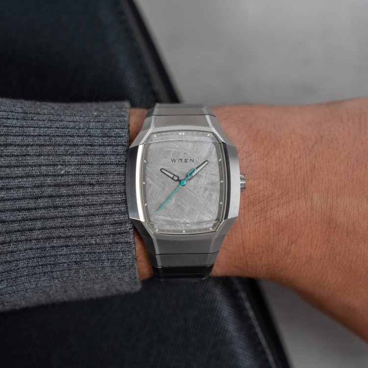 A person's arm wearing a silver Wren watch with a light gray, textured face and teal second hand.