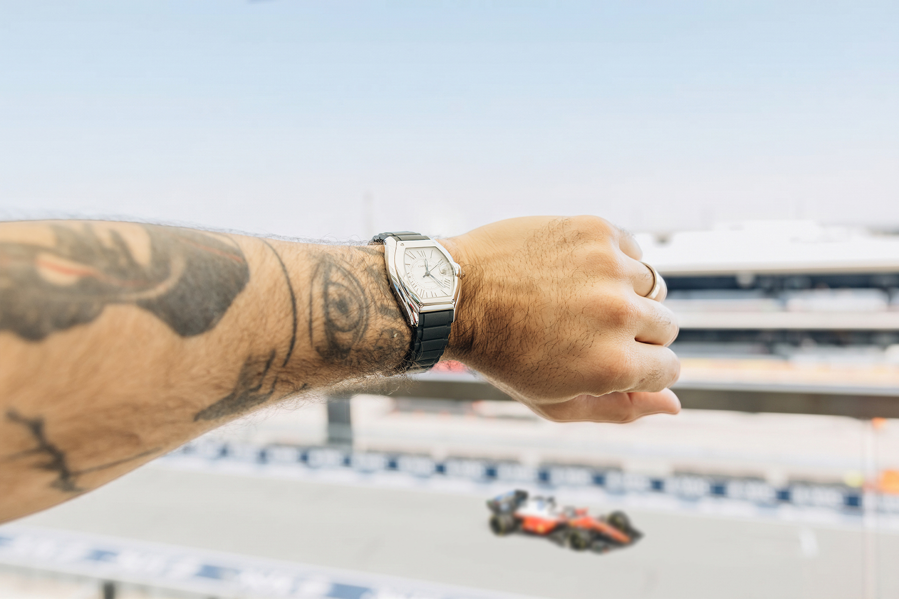 A tattooed arm wearing a silver watch with a black band, overlooking a blurred race track with a red F1 car.