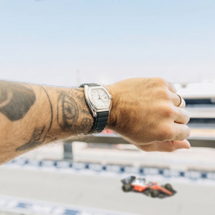 A tattooed arm wearing a silver watch with a black band, overlooking a blurred race track with a red F1 car.