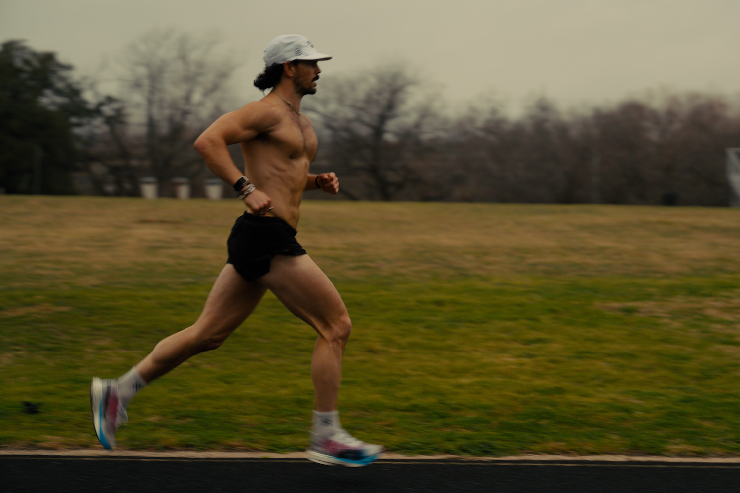 A shirtless man with a beard and white cap runs on a track, with a grassy field and trees in the background.