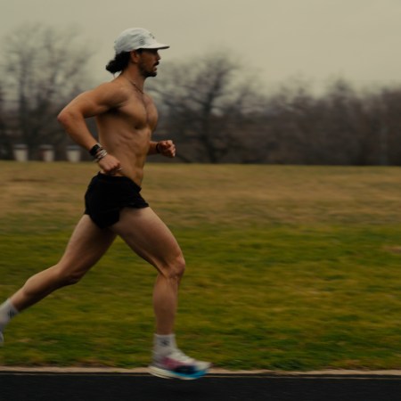 A shirtless man with a beard and white cap runs on a track, with a grassy field and trees in the background.