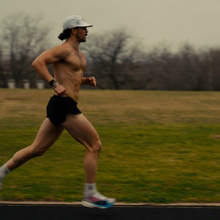 A shirtless man with a beard and white cap runs on a track, with a grassy field and trees in the background.