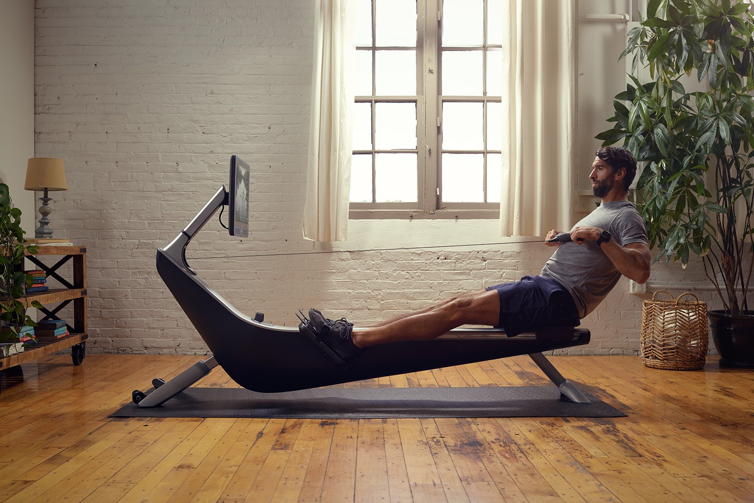 Man with a beard in a gray shirt and blue shorts rowing on a black rowing machine with a screen in a bright room.