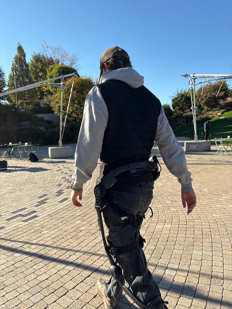 A person wearing a brown cap, gray hoodie, and black vest walks away from the camera, wearing an exoskeleton on their legs.