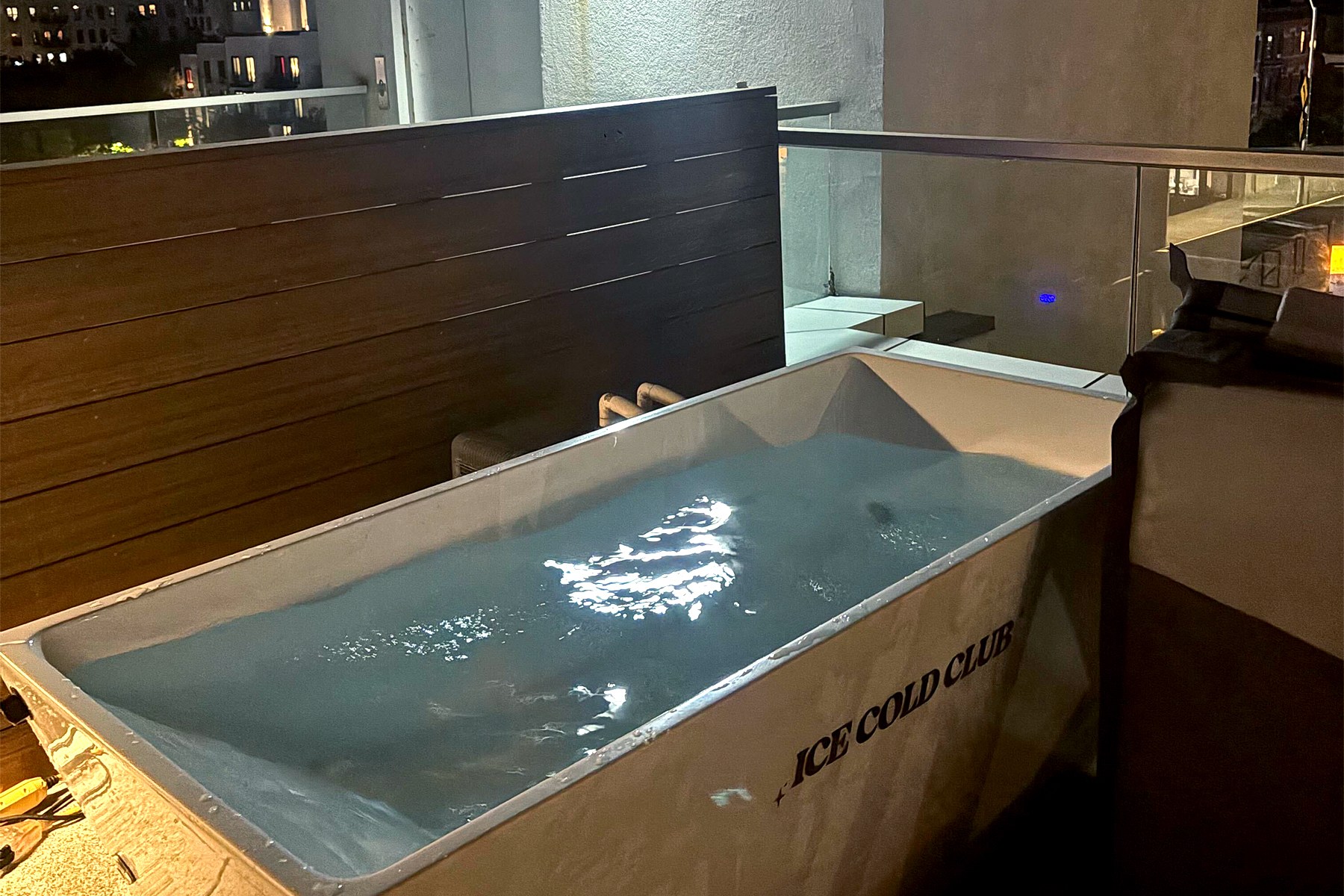 An "ICE COLD CLUB" ice bath filled with water on a balcony at night, with city lights in the background.