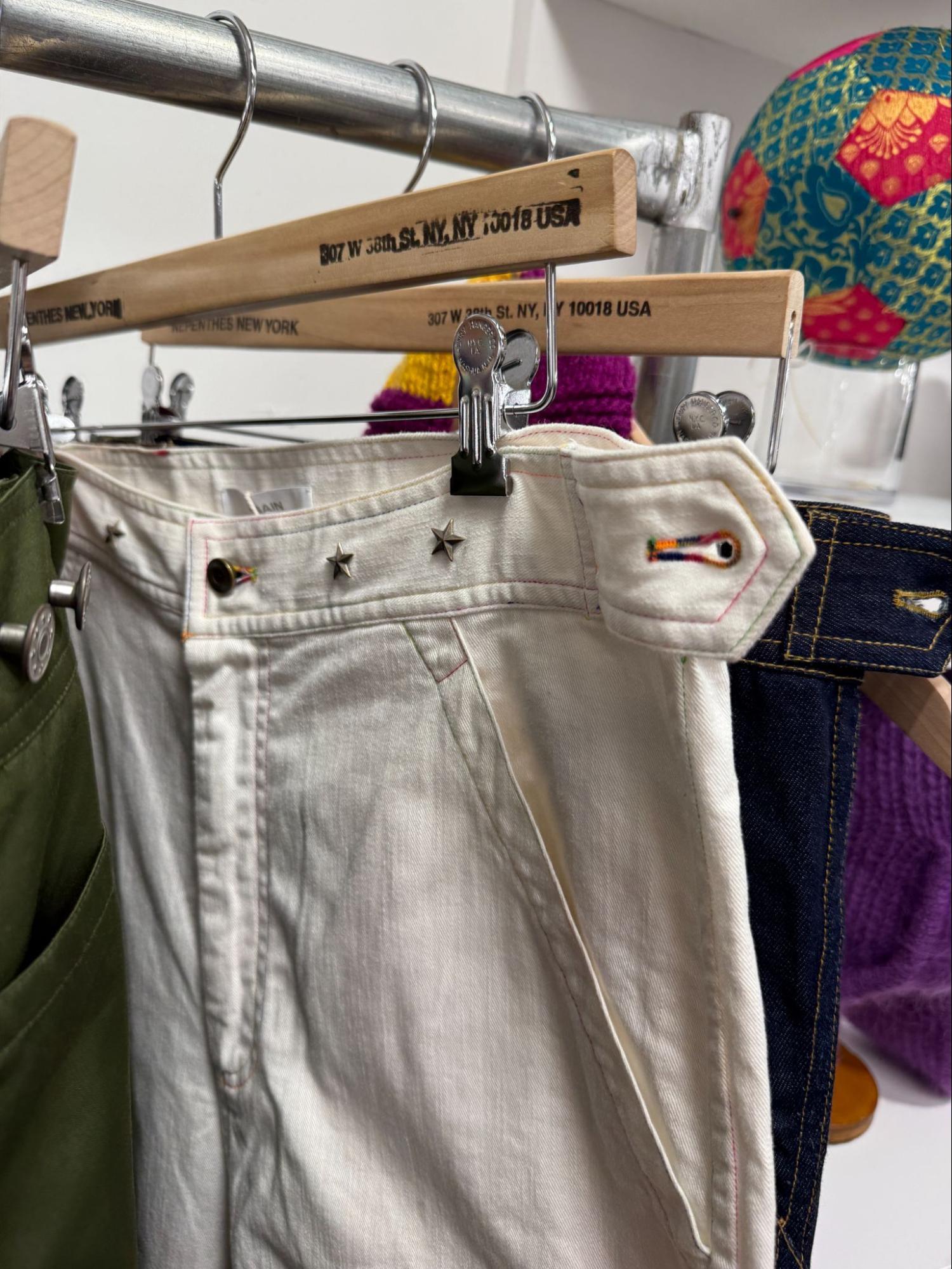 Close-up of white pants with star studs and colorful stitching, hanging on a rack with other clothes.