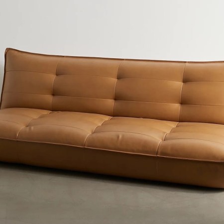 Tan leather futon with tufted cushions and a slightly curved backrest, on a gray floor against a white wall.