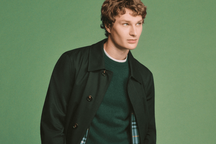 A man with curly brown hair and blue eyes, wearing a dark green jacket over a green sweater, looks right against a green background.