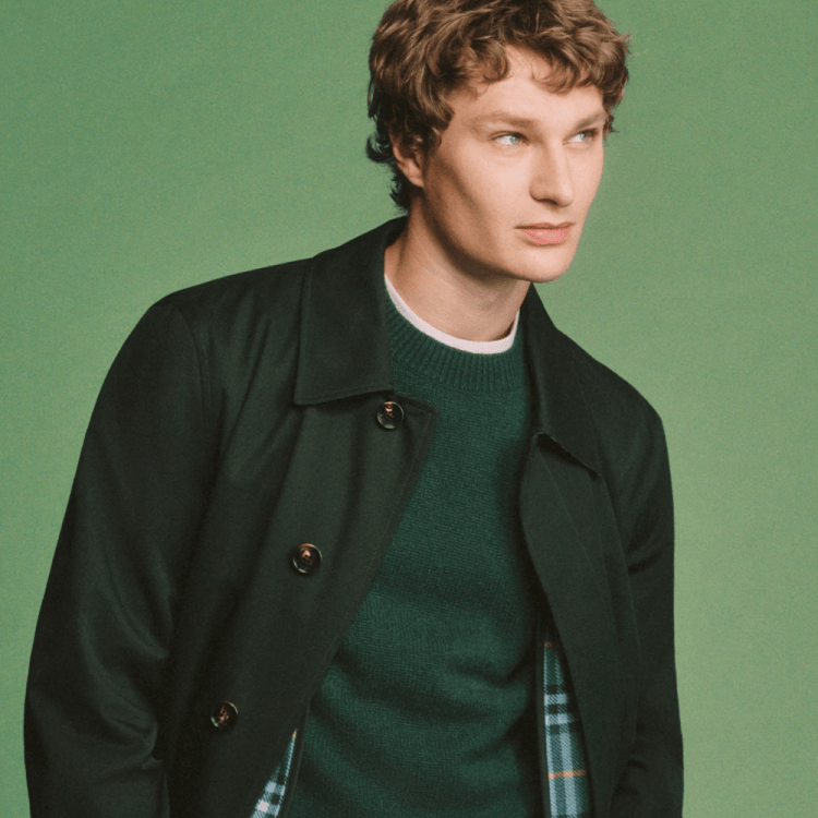 A man with curly brown hair and blue eyes, wearing a dark green jacket over a green sweater, looks right against a green background.