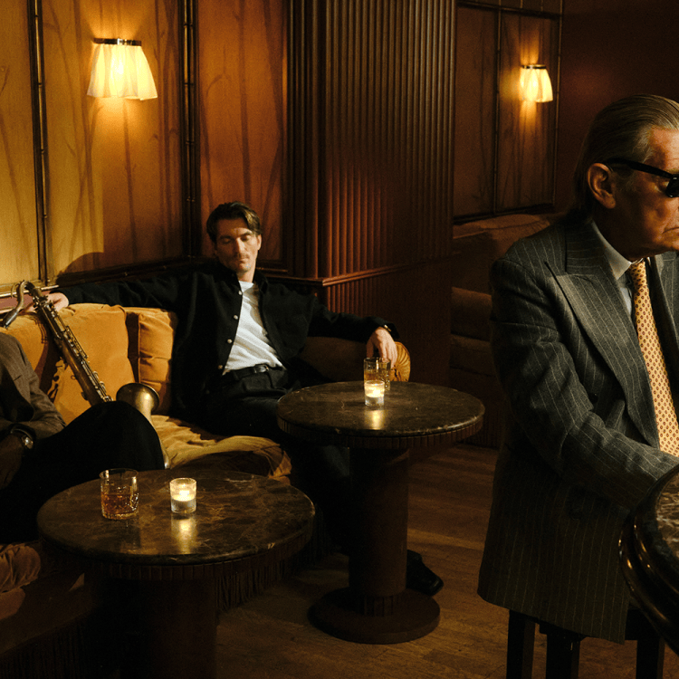 Three men in a dimly lit bar: two seated on a couch with a saxophone, one in sunglasses at a marble bar.