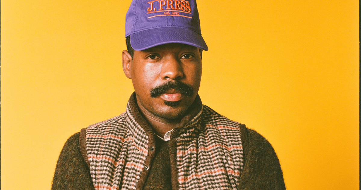 Man with a mustache wearing a purple J. Press cap, a brown sweater, and a plaid vest against a yellow background.