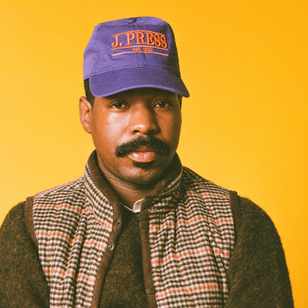 Man with a mustache wearing a purple J. Press cap, a brown sweater, and a plaid vest against a yellow background.