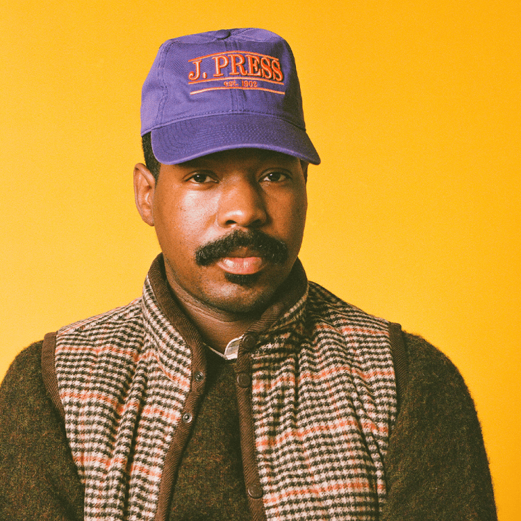 Man with a mustache wearing a purple J. Press cap, a brown sweater, and a plaid vest against a yellow background.